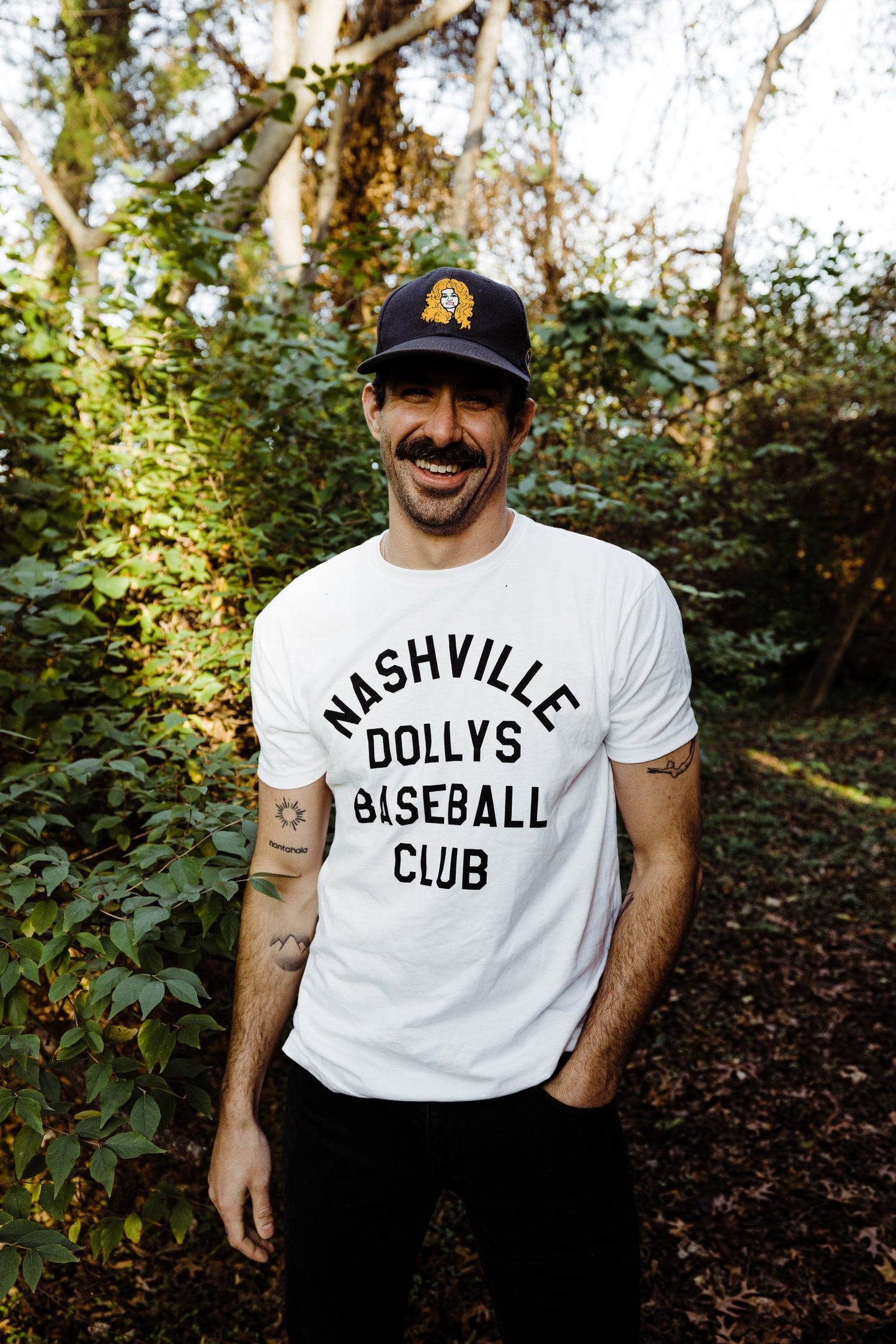 Model wearing the Nashville Dollys Baseball Club white t-shirt outdoors, paired with a navy cap.