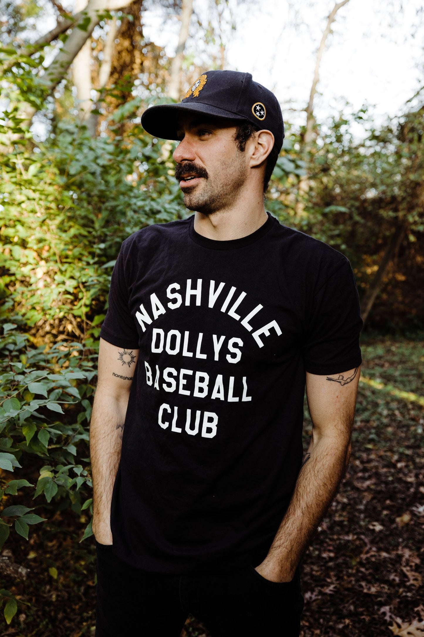 Man wearing a black t-shirt with white text 'Nashville Dollys Baseball Club' standing outdoors, surrounded by greenery.